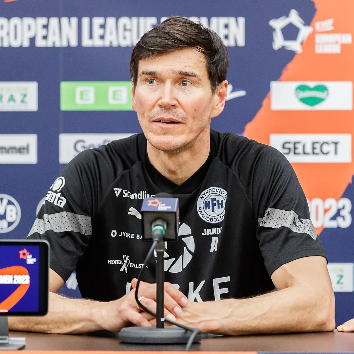 A man in a black Nykøbing Falster Handball shirt speaks at a post-match interview table.