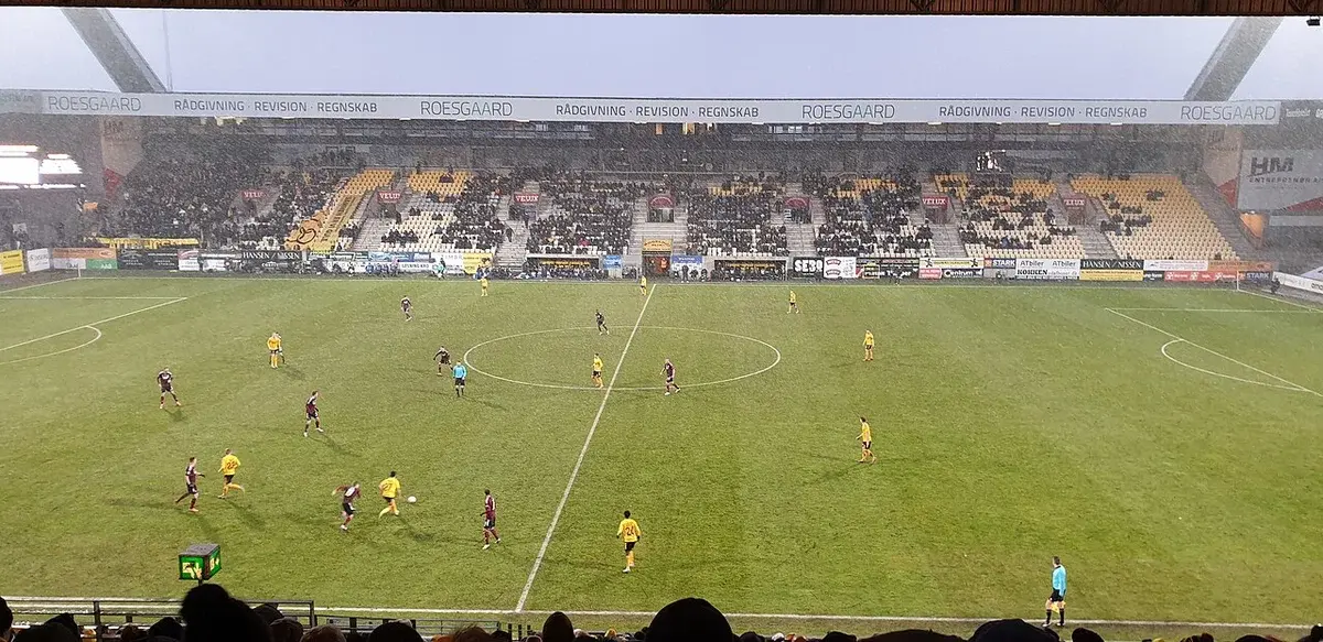 Wide shot of a football match in a stadium with players on the pitch and spectators in the stands.