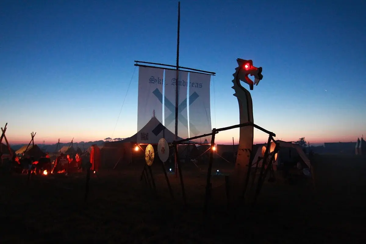 A scouting camp at dusk with tents, campfires, and a tall banner with a cross.