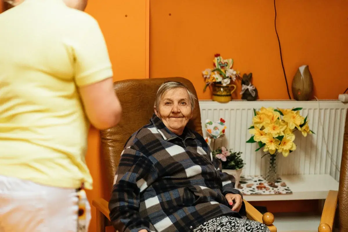 An elderly woman sits in an armchair while a caregiver stands nearby in a care home room.
